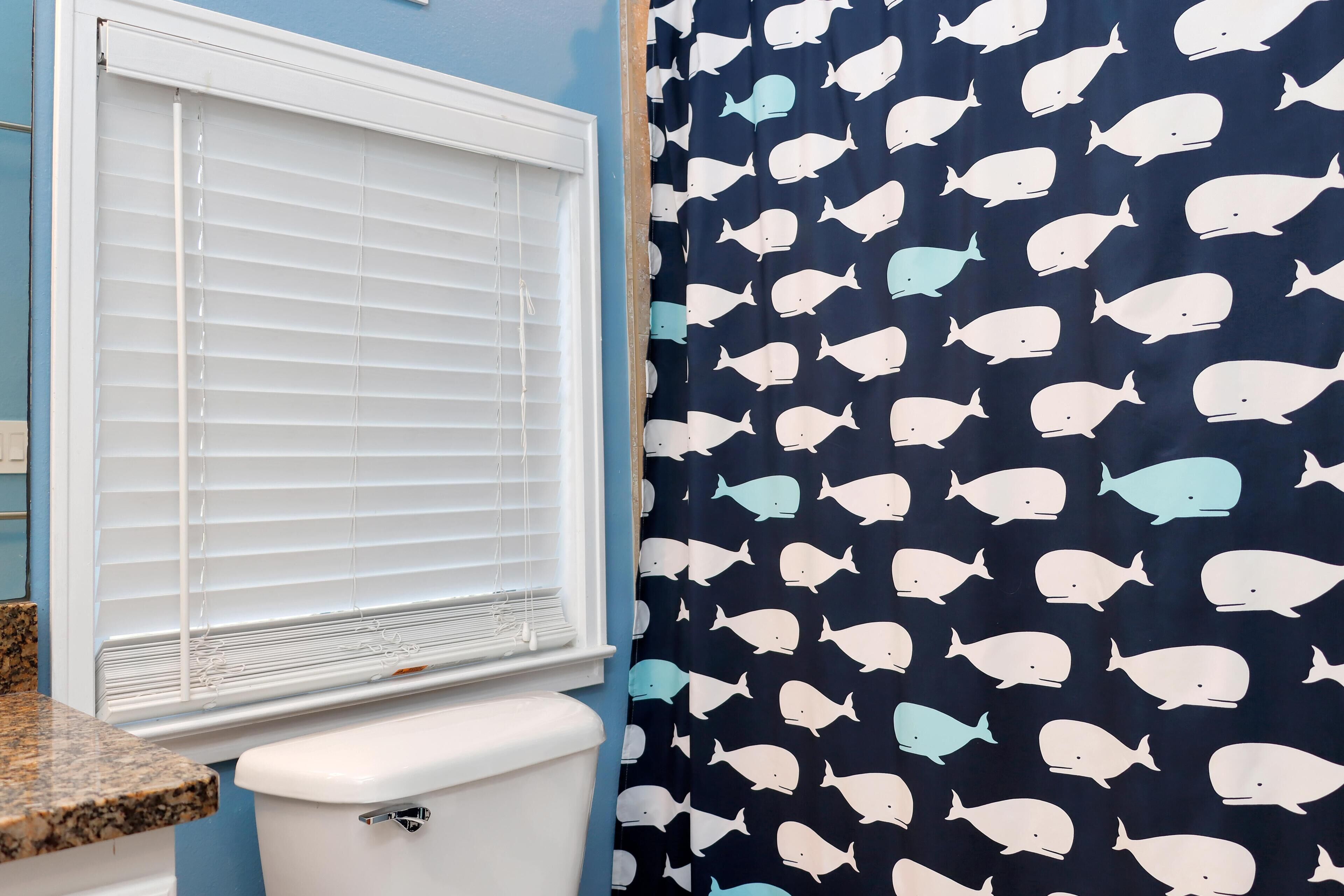 Une salle de bain avec des toilettes, une fenêtre avec des stores et un rideau de douche orné d’un motif de baleine.