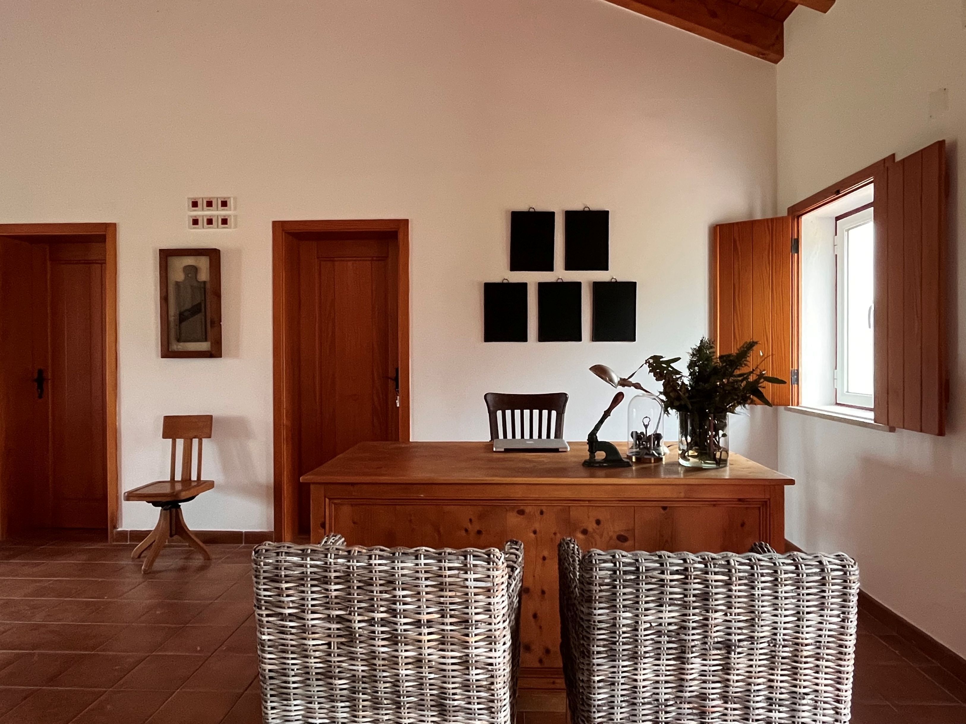A dining area with a wooden table, wicker chairs, a small desk, and a window with wooden shutters.
