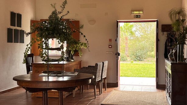 A dining area with a round table, chairs, a buffet counter, and a view of a garden through an open door. - Monte da Estrada - Turismo Rural (Zambujeira do Mar)