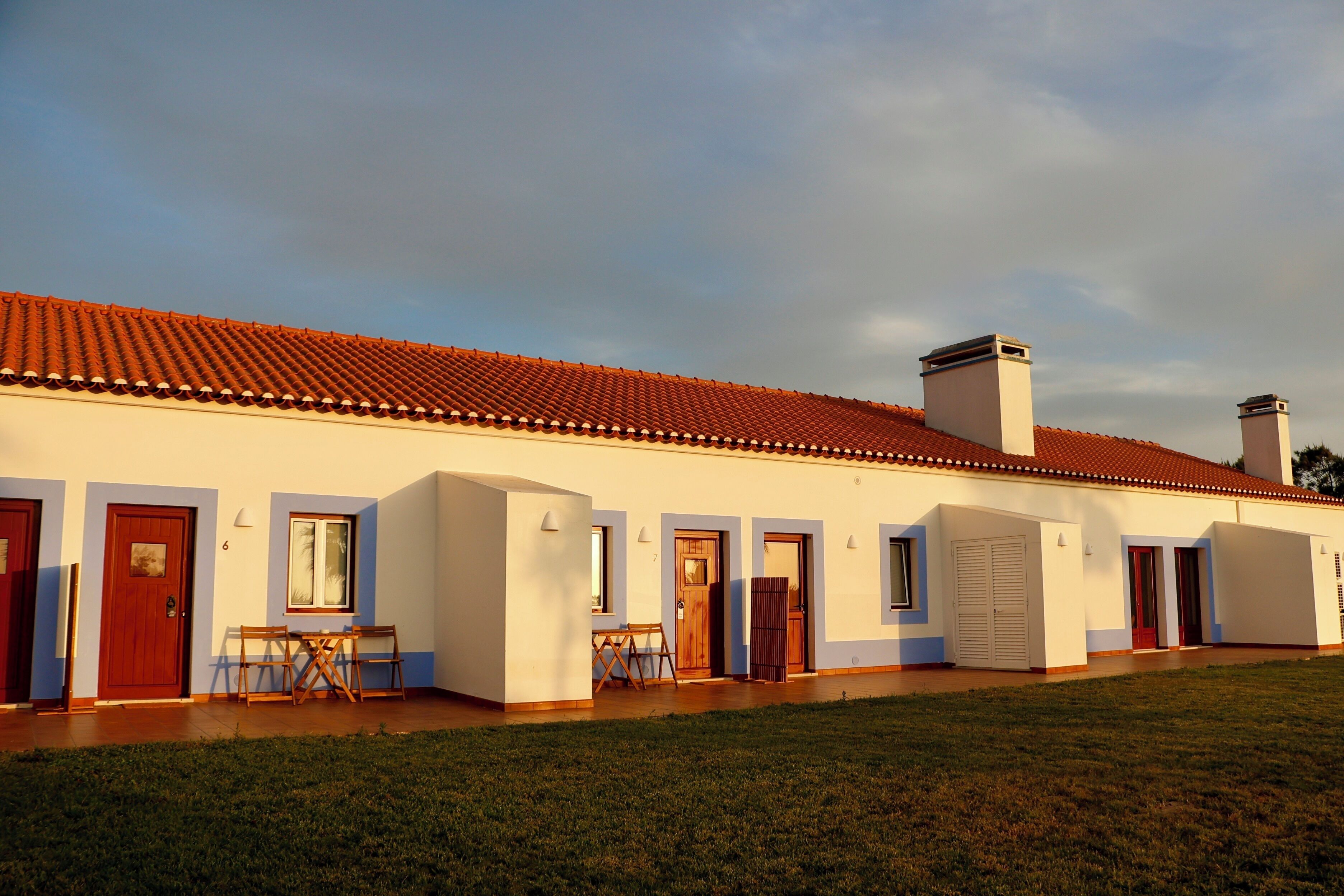 Un bâtiment à deux étages, doté d’un toit recouvert de tuiles rouges, de plusieurs portes et d’un petit patio aménagé avec des chaises.