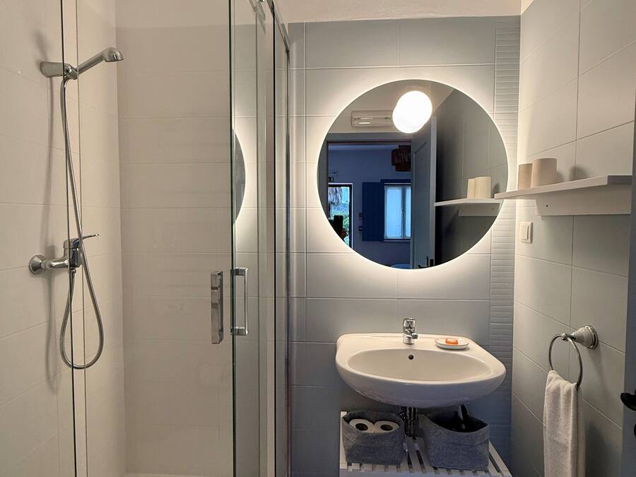 Un baño moderno con ducha de vidrio, lavamanos blanco y espejo redondo con luz.