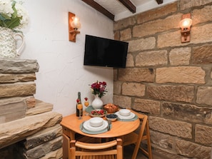 A stone wall interior with a flat-screen TV mounted above a wooden table set for two, featuring plates, glasses, and a vase with flowers. - Bishop's Cottage (Whitby)
