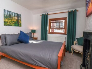 A bedroom with a bed, a chair, a fireplace, a window with curtains, a lamp, and a painting on the wall. - Bishop's Cottage (Whitby)