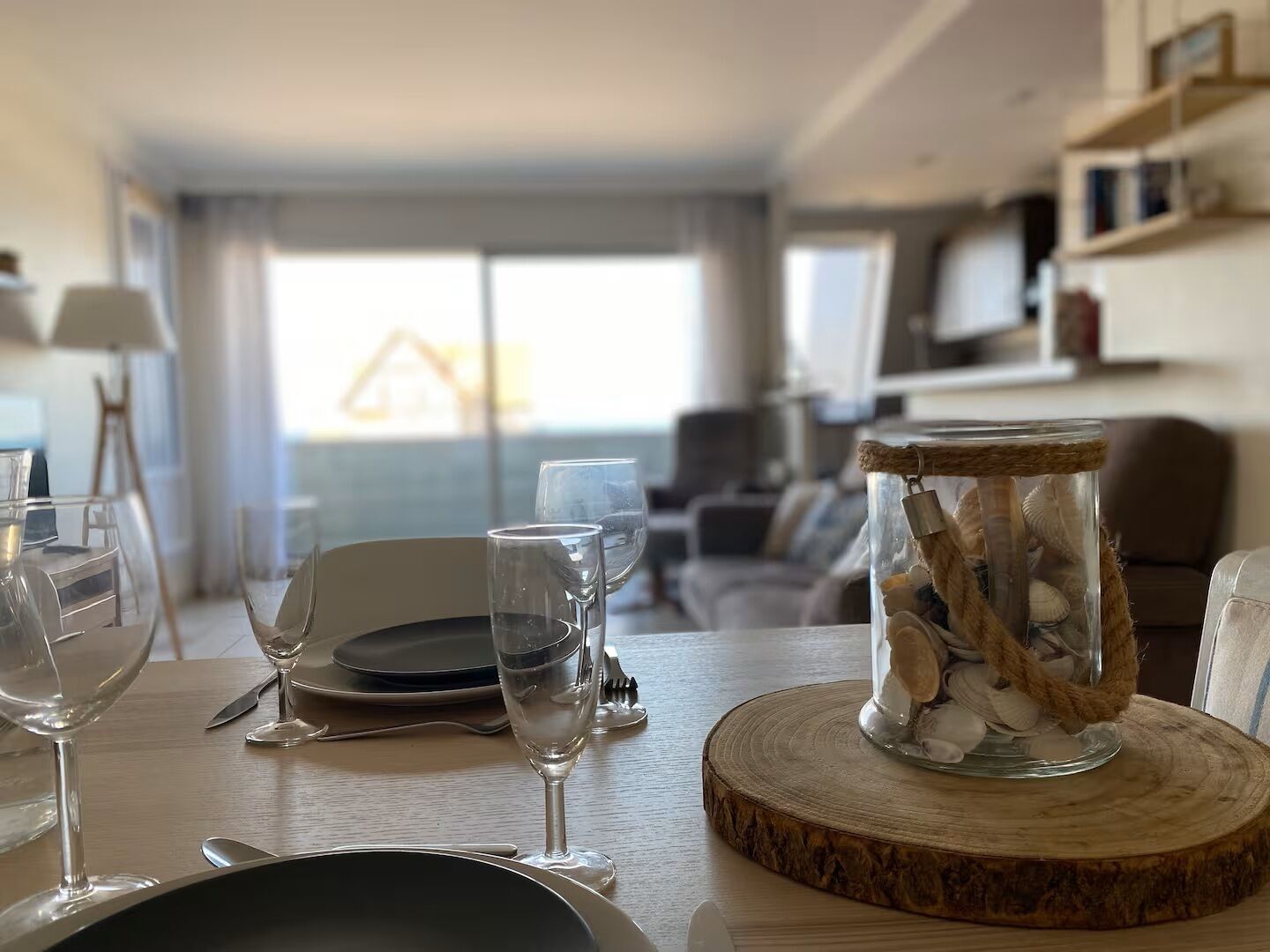 Une table à manger dressée avec des assiettes, des verres et des couverts, un bocal en verre rempli de coquillages et un sous-verre en bois.