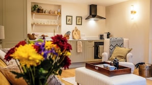 A living room with a sofa, a coffee table with a tray, a kitchen area with shelves, and a wall-mounted light. - Sycamore Cottage Banham Cottages (Bourton-on-the-Water)