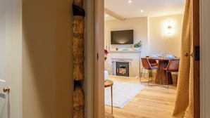 A room with a fireplace, a dining table with chairs, a television mounted on the wall, and a patterned curtain. - Sycamore Cottage Banham Cottages (Bourton-on-the-Water)