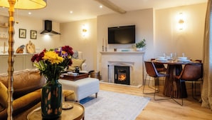 A living room with a sofa, a coffee table, a vase with flowers, a fireplace, a dining table with chairs, and a flat-screen TV mounted above the fireplace. - Sycamore Cottage Banham Cottages (Bourton-on-the-Water)