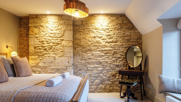 A bedroom with a stone wall, a bed with a rolled-up blanket, a bedside table with a lamp, and a mirror. - Sycamore Cottage Banham Cottages (Bourton-on-the-Water)