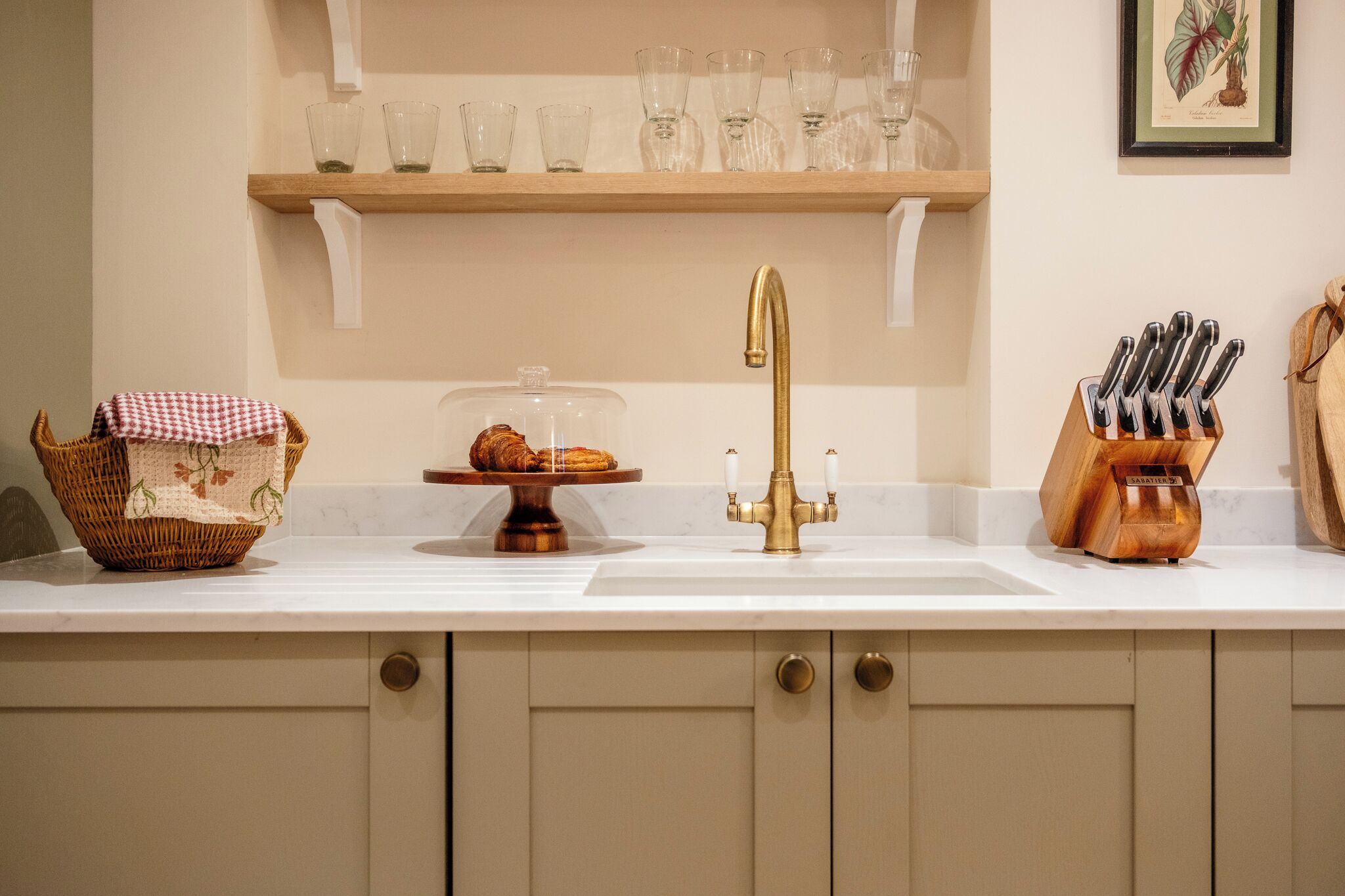 A kitchen with a white countertop, a gold faucet, a basket with a cloth, a cake on a stand, and a knife block.