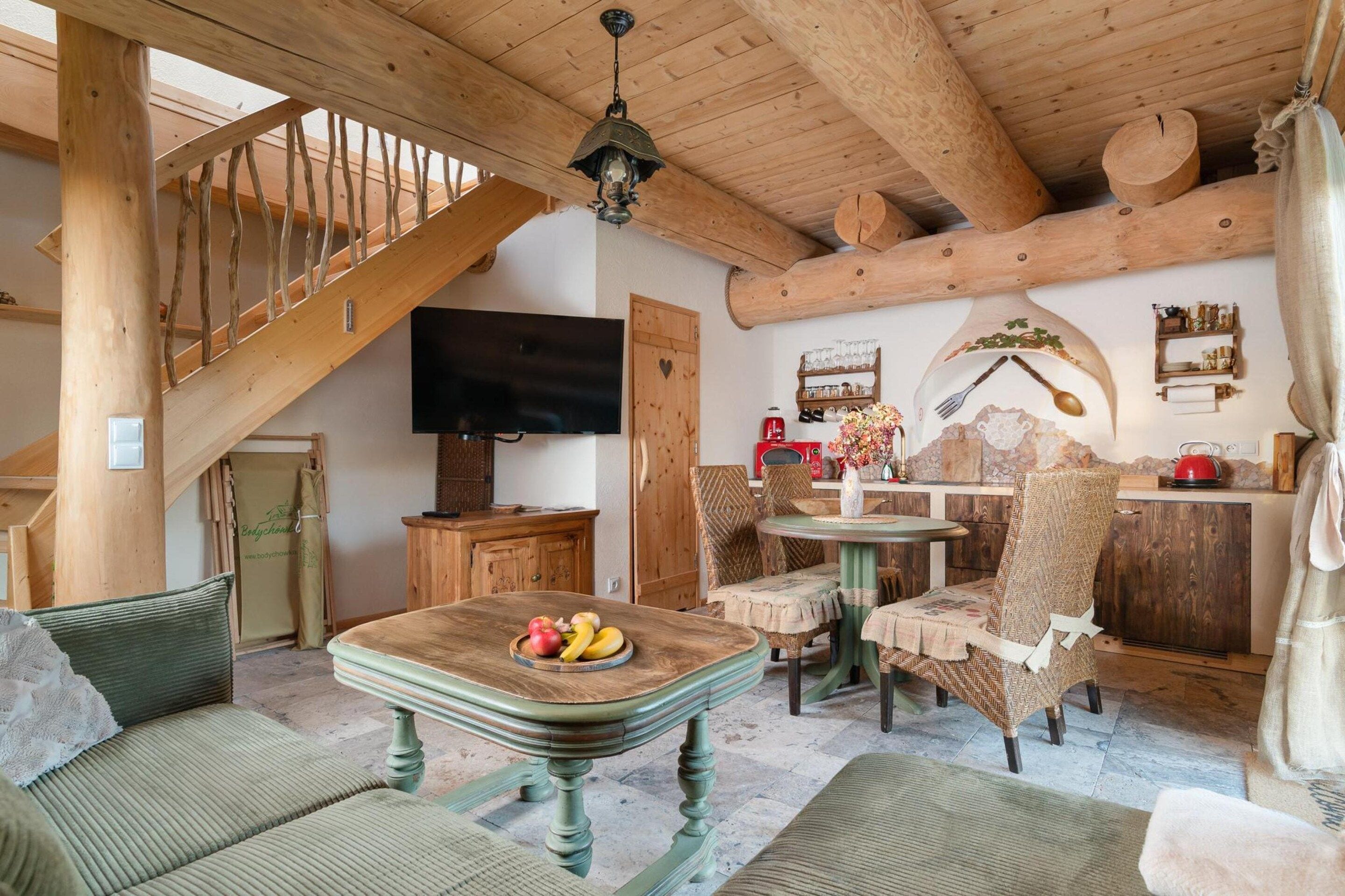 A rustic interior with a wooden staircase, a flat-screen TV, a dining area with wicker chairs, and a table with fruit.