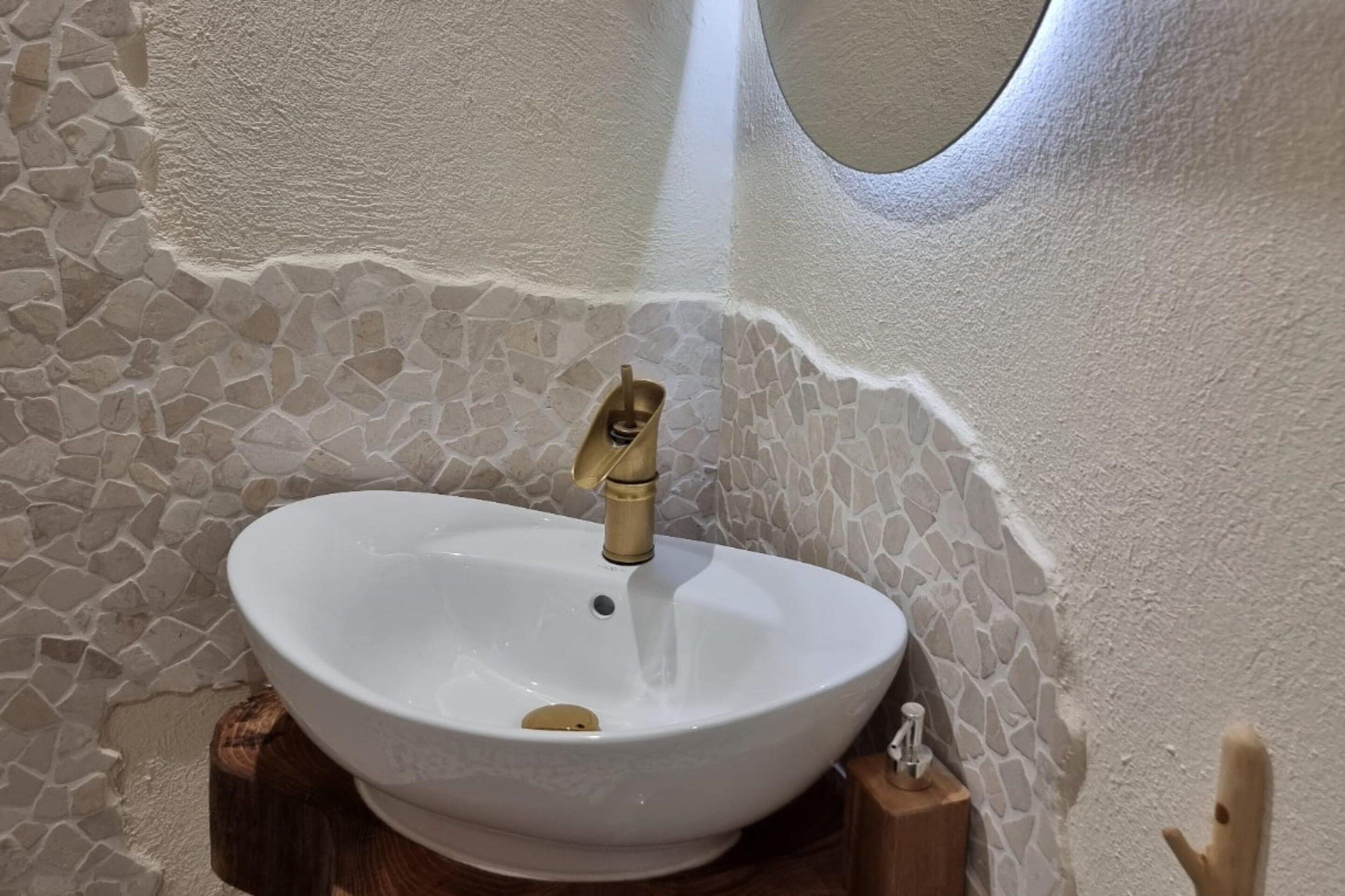 Une salle de bain avec un lavabo blanc, un mur décoré d’une mosaïque de pierres et un socle en bois.