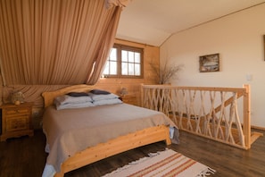 A wooden bed with a headboard, a nightstand, a bedside table, a window with curtains, a wooden railing, and a painting on the wall. - Comfortable Apartment Very Close to the Beach (gmina Będzino)