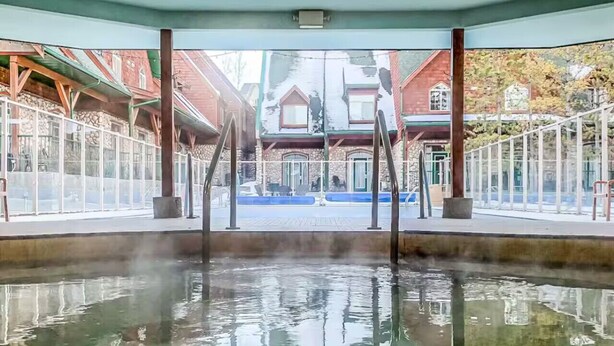 An indoor swimming pool with clear water, wooden deck, and a view of a building with a stone facade. - Mountain Chic | 2BR Family Escape | Hot Tub + Pool (Canmore)