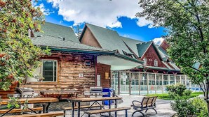 A rustic cabin-style building with outdoor seating, a barbecue grill, and a glass-enclosed area. - Mountain Chic | 2BR Family Escape | Hot Tub + Pool (Canmore)