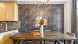 A dining area with a wooden table, chairs, a vase with dried leaves, and a modern kitchen with a stone backsplash. - Mountain Chic | 2BR Family Escape | Hot Tub + Pool (Canmore)