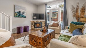 A living room with a stone fireplace, a television, and a view through glass doors. - Mountain Chic | 2BR Family Escape | Hot Tub + Pool (Canmore)