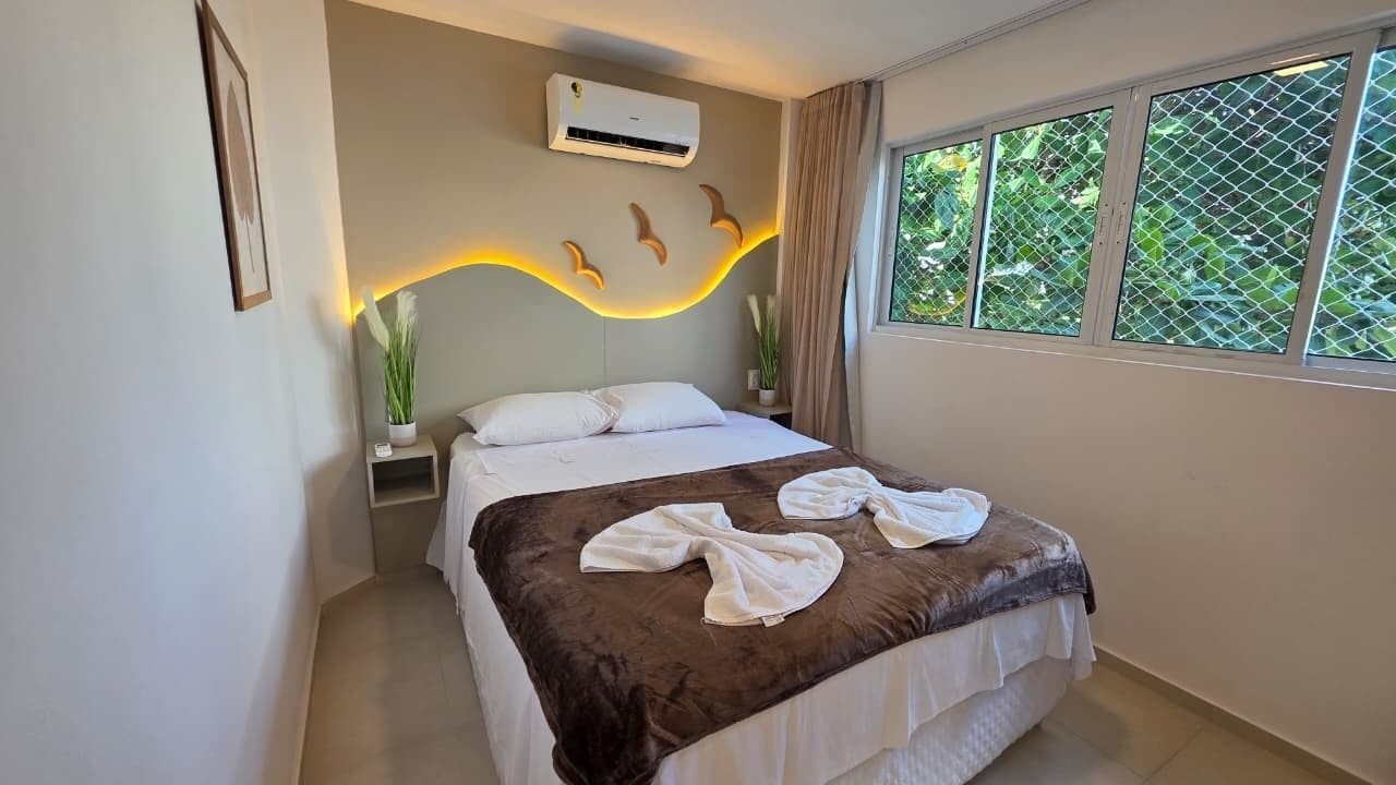 A hotel room with a bed, an air conditioner, a window with a view of greenery, and a wall-mounted light fixture.
