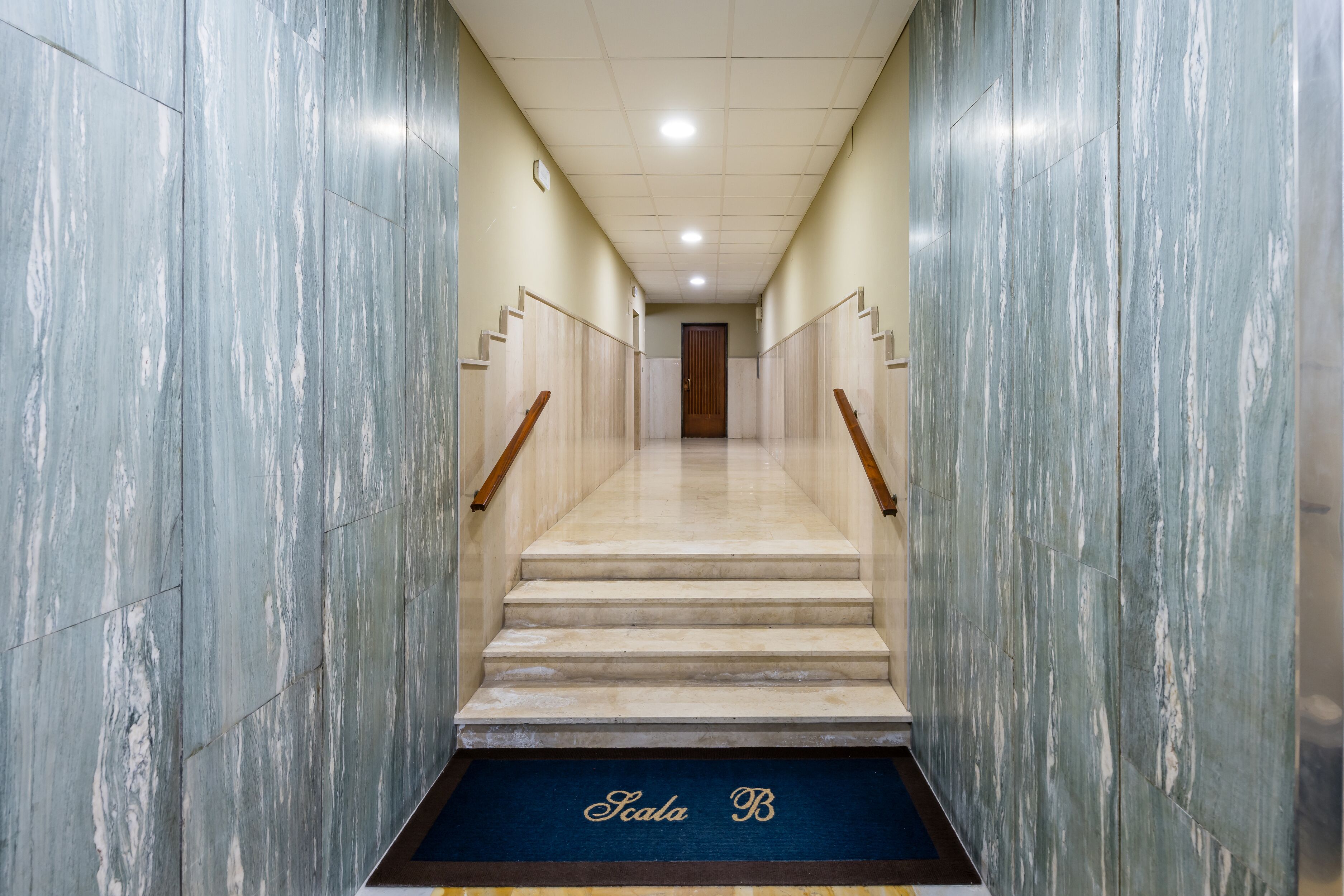 A hallway with marble walls, a wooden handrail, and a carpeted landing with 'Scala B' text.