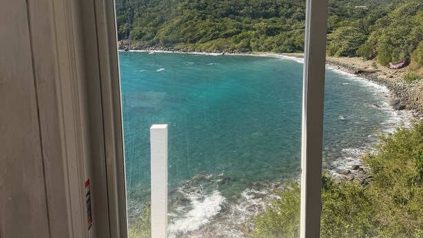 Janela com vista para a paisagem costeira, incluindo uma praia e uma colina arborizada.