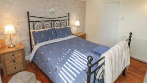 A bedroom with a bed, two bedside tables, a nightstand, a lamp, a radiator, and a door. - Puffin's Nest (Bridlington)