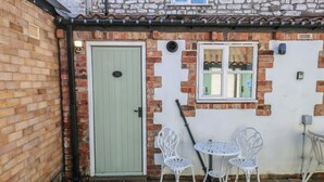 Un bâtiment en briques avec une porte verte, une fenêtre et deux chaises en métal blanc.