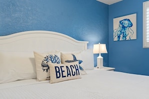 A bedroom with a white bed, blue walls, and a single white nightstand with a lamp. - Steps to Beach! Ground-Floor Condo w/ Pool Sleep 4 (Panama City Beach)