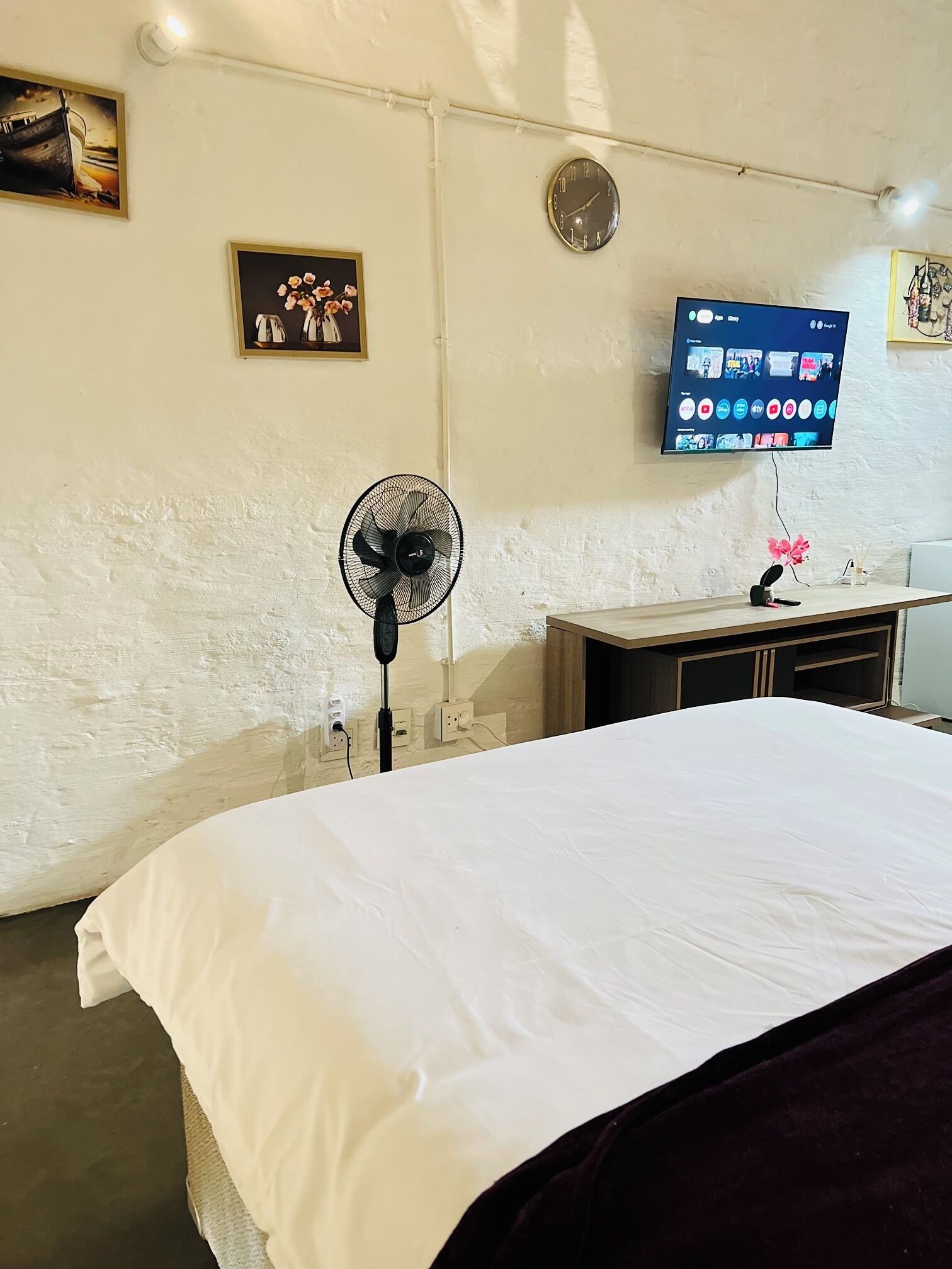A bedroom with a bed, a wall-mounted TV, a fan, and a clock.