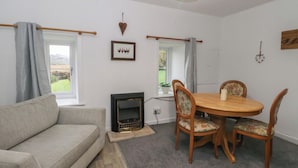 A cozy living room with a sofa, a dining table with chairs, a fireplace, and a view through a window. - Holly Cottage (Inveraray)