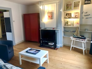 A living room with a white coffee table, a red cabinet, a TV stand, and a bookshelf with various items. - L'orchidée: Your Apartment in the Heart of Honfleur (Honfleur)