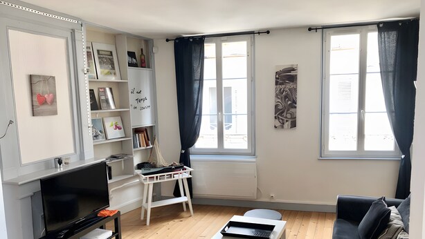 Uma sala de estar com televisão, uma mesa branca, um sofá preto, uma estante de livros e uma janela com cortinas.