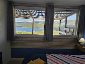 Ein Schlafzimmer mit großem Fenster, einem Bett, einem Nachttisch und Blick auf einen See und Hügel.