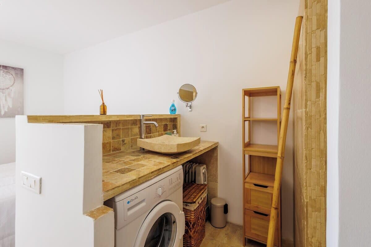 A compact bathroom with a washing machine, a sink with a mirror, and a wooden shelf.