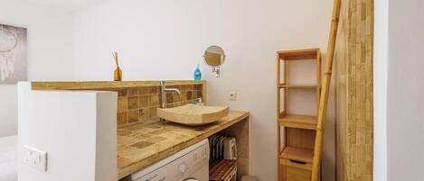 A compact bathroom with a washing machine, a sink with a mirror, and a wooden shelf.