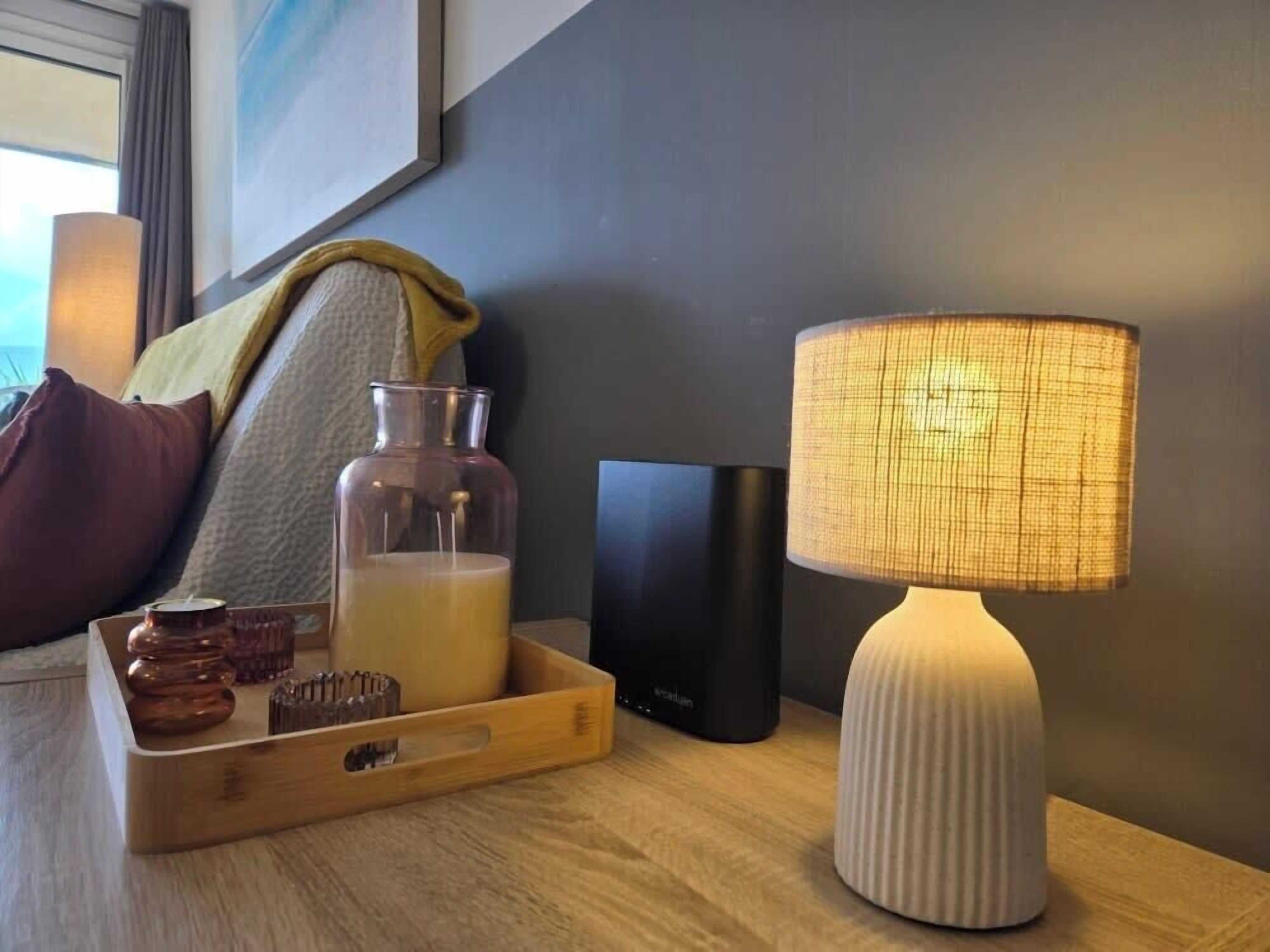 A wooden bedside table with a white lamp, a glass bottle, a black box, and a wooden tray with a candle and a small container.