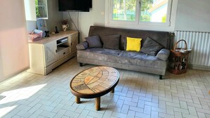 Ein Wohnzimmer mit grauem Sofa, einem Couchtisch, einem an der Wand befestigten Fernseher und einem Fenster mit Blick ins Grüne.