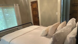 Uma cama com roupa de cama e almofadas brancas, uma janela com cortinas e uma porta de madeira.