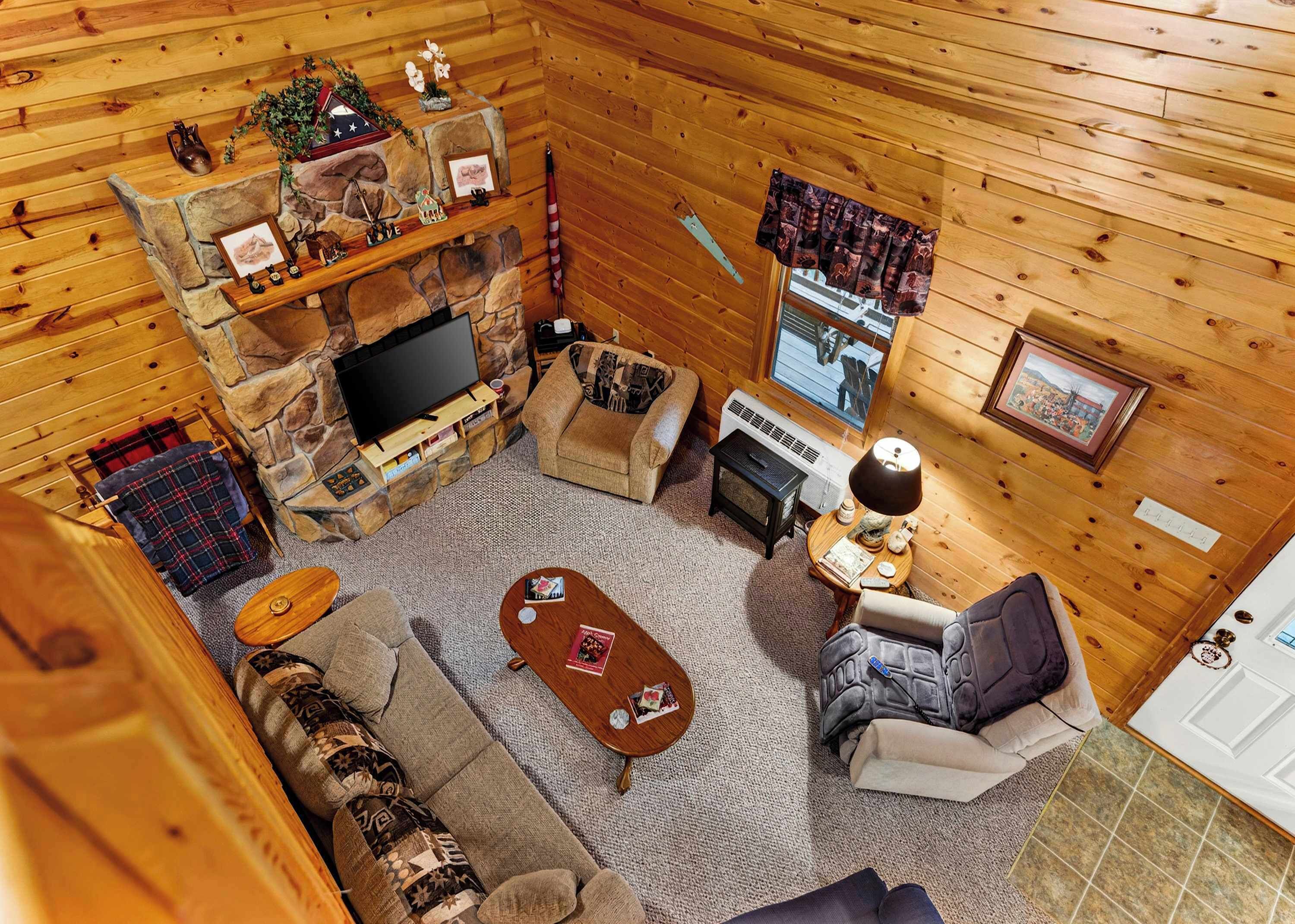 A cozy cabin interior with a stone fireplace, a television, a sofa, a coffee table, and a window with curtains.