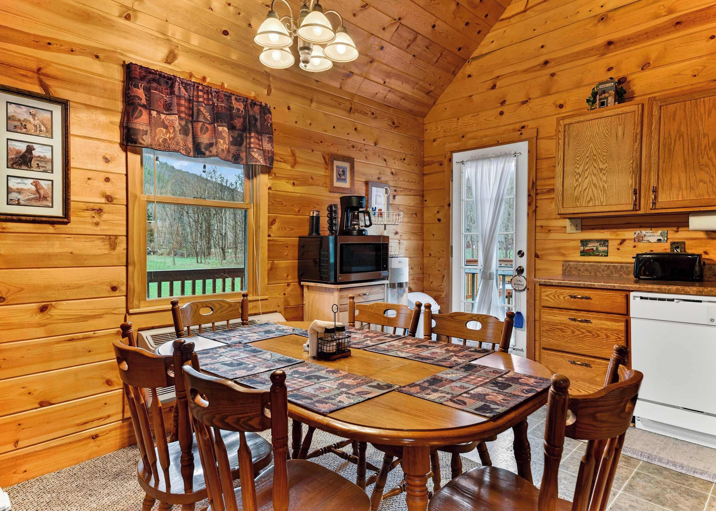 A wooden cabin interior with a dining table, chairs, a kitchen area, and a window with a view of trees.