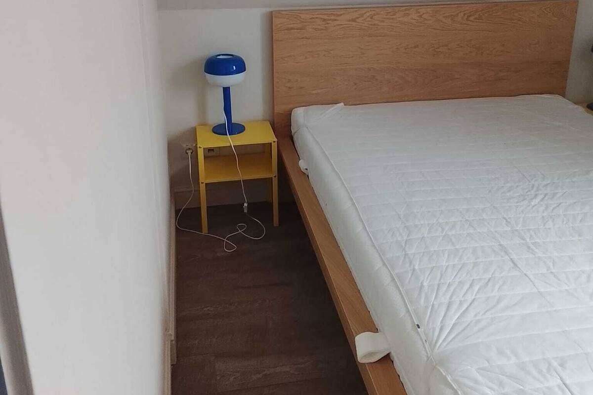 A single bed with a wooden headboard, a bedside table with a lamp, and a power outlet on the wall.