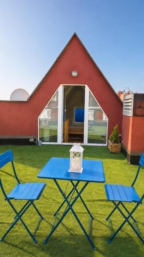 A red-roofed building with a triangular window, a small table with a white lantern, and blue folding chairs on a green lawn. - Dzien Dobry Hostel (Gdańsk)