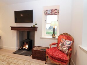 Un salon doté d’une cheminée, d’un téléviseur fixé au mur, d’une fenêtre ornée d’un rideau à motifs et d’un fauteuil rouge recouvert de tissu, agrémenté d’un coussin.