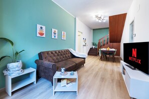 Un salon avec un canapé marron, une table basse, un téléviseur affichant le logo de Netflix et un escalier menant à une salle à manger.