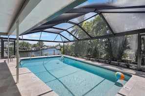 An indoor swimming pool with a view of a body of water and palm trees, surrounded by a covered area with a metal frame and glass panels. - 737 Nautilus Court (Marco Island)