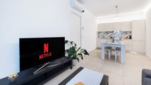 A modern living room with a flat-screen TV displaying the Netflix logo, a dining area with a white table and chairs, a kitchen with marble countertops, and a potted plant. - Cattolica Modern Beach - Riviera Collection (Cattolica)