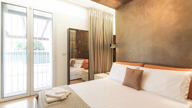Une chambre à coucher moderne avec un grand lit, une porte-fenêtre coulissante donnant sur un balcon, un miroir et une palette de couleurs neutres.