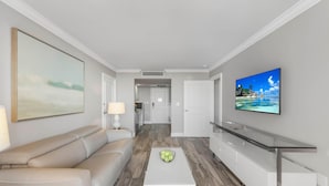 Uma sala de estar moderna com sofá, mesa de centro, TV e área de cozinha ao fundo.