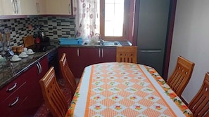 A kitchen with a checkered tablecloth, wooden chairs, a refrigerator, and a window with curtains.