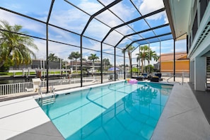 An indoor swimming pool with a glass enclosure, surrounded by a white fence, and palm trees in the background. - 1672 Barbarosa Court (Marco Island)