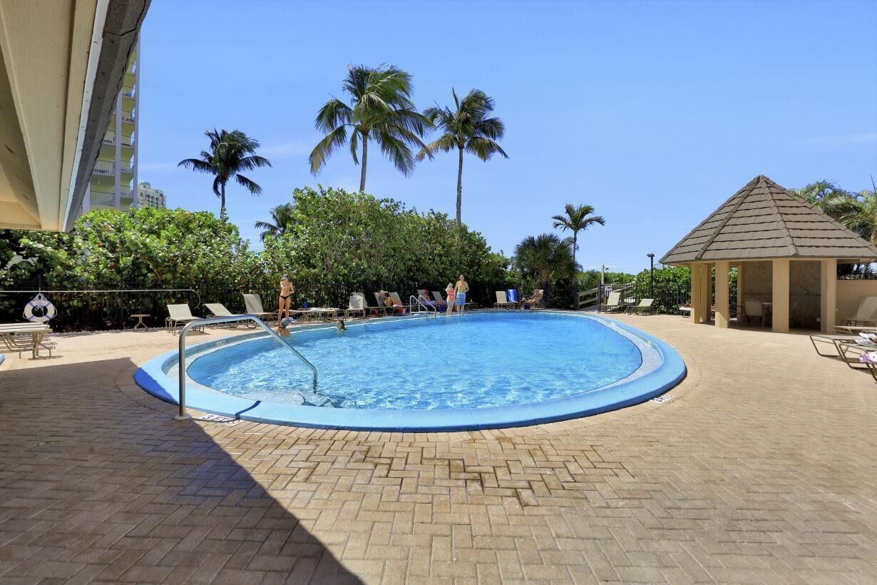 A swimming pool with lounge chairs and a gazebo, surrounded by palm trees and a brick patio.