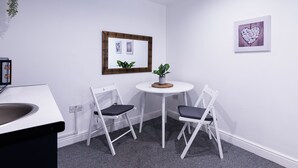 Une petite pièce avec un bureau blanc, deux chaises blanches, une table ronde ornée d’une plante et deux tableaux encadrés accrochés au mur.